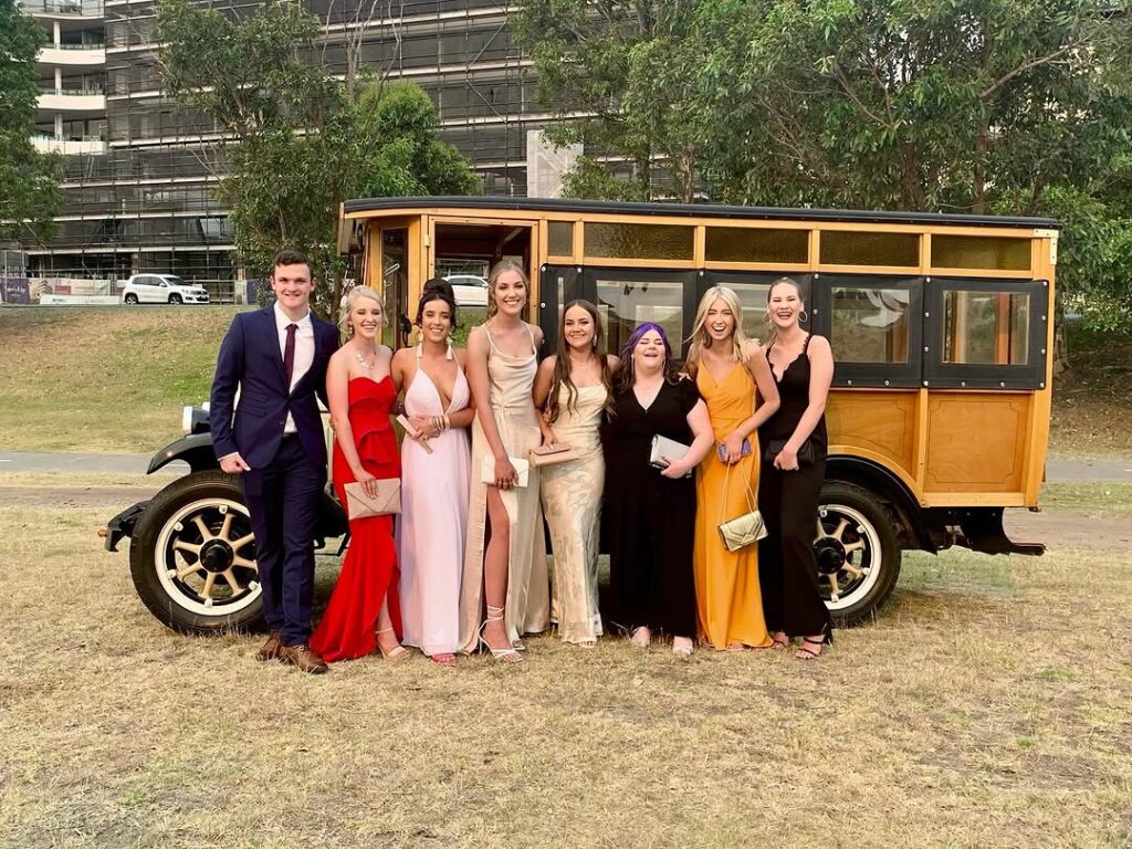 School formal group posing beside a vintage woody bus parked on dry grass.