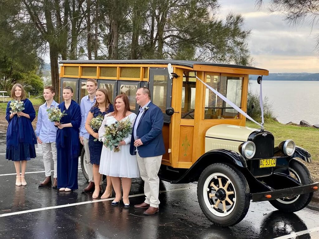 Wedding group posing beside a decorated 1927 Chev Woodie bus on wet pavement near the water.