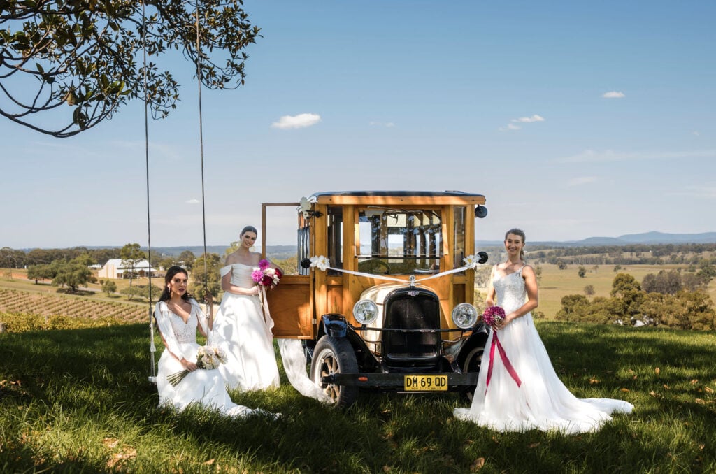 Three brides in white dresses posing with a decorated vintage woody bus in a vineyard setting.