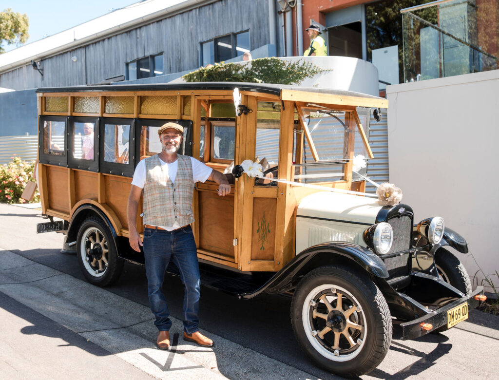 Hunter Woodies driver standing beside a decorated vintage woody bus parked on a paved street.